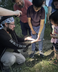 Caleb Hinojos, kneeling on the ground outside in early twilight, showing a group of children and adults a small insect on his hand while an unseen person shines a flashlight on the insect.